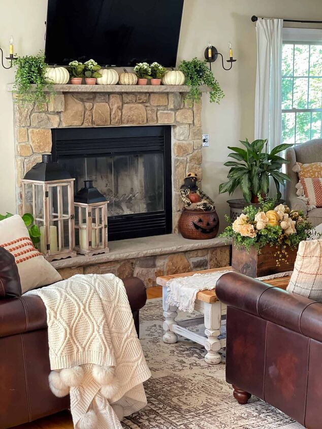 7 easy ways to make faux flowers look real, rustic farmhouse living room decorated for fall with faux pumpkins and greenery with fireplace and tv over the mantel with dark leather sofas and cozy throw blankets with neutral fall decorating ideas